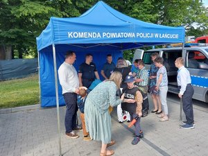 Policyjne stoisko znajdujące się pod namiotem z napisem: Komenda Powiatowej Policji w Łańcucie. Obok stoiska stoi radiowóz z otwarty drzwiami. Przy stoisku stoją uczestnicy pikniku, którzy rozmawiają z policjantem. Obok nich chłopiec przymierza policyjne umundurowanie.