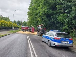 Ciągnik rolniczy znajdujący się na prawym poboczu ul. Sikorskiego. Za nim znajdują się dwie przyczepy stojące na całej szerokości jezdni. Jedna leży na boku. Wokół nich wysypane są ziarna kukurydzy.