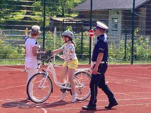 Uczestniczka egzaminu na kartę rowerową stoi przed linią z napisem STOP. Obok niej stoi policjant oraz nauczycielka.