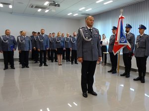 Na środku sali stoi odznaczany policjant. Za nim, w trzech rzędach, stoją policjanci, którzy otrzymali w tym roku awans na wyższy stopień służbowy.