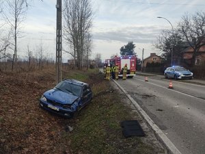 Obok jezdni, w przydrożnym rowie, znajduje się samochód marki Peugeot. Samochód ma uszkodzony przód, ugiętą pokrywę silnika oraz dach.