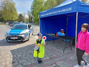 Chłopiec ubrany w policyjne umundurowanie pozuje przed policyjnym stoiskiem i oznakowanym radiowozem.