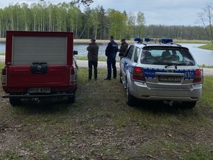 Policjant wraz z dwoma strażnikami leśnymi pełni służbę na terenie leśnym, w pobliżu stawu.