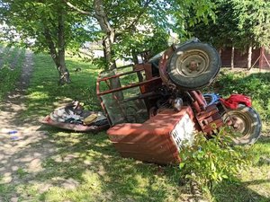 Leżący na prawym boku ciągnik rolniczy. Za nim droga oraz skarpa, z której pojazd zjechał i się przewrócił.