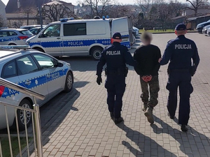 Dwóch umundurowanych policjantów prowadzi mężczyznę z założonym kajdankami na rękach.