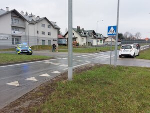 Ul. Armii Krajowej w Łańcucie z przejściem dla pieszych obok którego, na trawie leży przewrócona hulajnoga. Po drugiej stornie ulicy stoi częściowo na trawie samochód marki Chevrolet .
