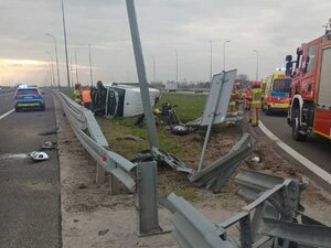 Miejsce wypadku. Na boku poza barierkami energochłonnymi leży uszkodzony pojazd marki Mercedes Sprinter. Na jezdni stoi oznakowany radiowóz.